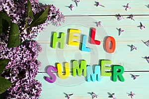 On a wooden background there is an inscription in multicolored letters hello summer surrounded by lilac flowers