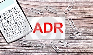 On a wooden background, a white calculator, white paper clips and a white card with the text ADR Alternative Dispute Resolution.