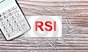 On a wooden background, a white calculator, white paper clips and a white card with the text RSI Relative Strength Index. View