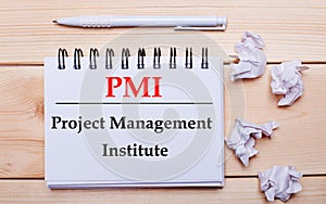 On a wooden background, a white notebook with the inscription PMI Project Management Institute, a white pen and crumpled white