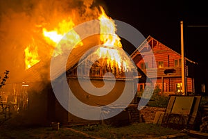 Wooden barn burning at night. High orange fire flames, dense smoke from under tiled roof on dark sky, trees silhouettes and reside