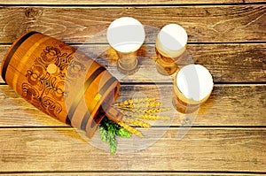 A wooden barrel and three glass glasses of light beer stand on a wooden table, next to them are hops and ears of barley