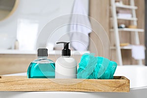 Wooden bath tray with bottles of shower gels and mesh pouf on tub indoors