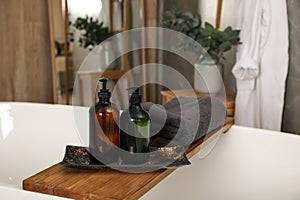 Wooden bath tray with bottles of shower gels and towel on tub indoors, space for text