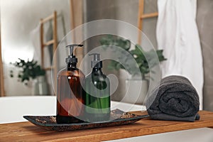 Wooden bath tray with bottles of shower gels and towel on tub indoors, space for text