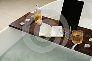 Wooden bath tray with candles, tablet, book and glass of drink on tub, closeup