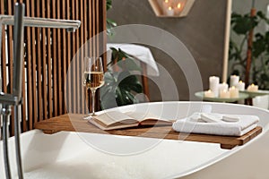 Wooden bath tray with glass of wine, open book, massage stones and towel on tub indoors. Relaxing atmosphere