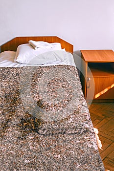 Wooden bed and bedside table next to it. The bed is covered with a brown bedspread and clean linen: white sheets, a pillow, and a