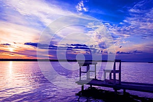 Wooden bench or chairs and table on a wood dock facing a calm lake at sunset. Sunset time on river. Orange sunset landscape with p