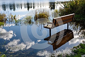 Wooden bench on the flooded shore of Larson Lake, reflections of blue sky and clouds