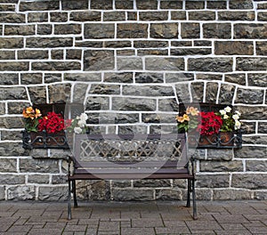 Wooden bench in front of old gray stone wall with two small windows decorated with colorful flowers