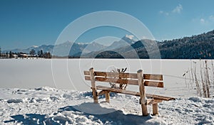 Wooden bench by the frozen Weissensee lake before the mountains on a sunny winter day