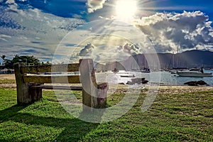 Wooden bench and stone by the sea at incredible sunset with sun rays passing between clouds and boats in the background in Ilhabel