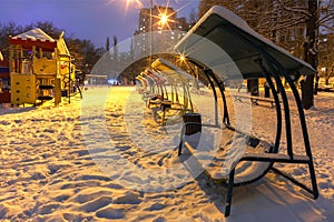 Wooden benches and a playground in the winter city evening park are covered with snow against the background of blue twilight