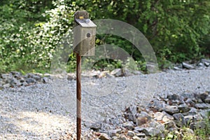 Wooden bird box house with hole and door on rusty post with gray gravel rock path and green plants in background