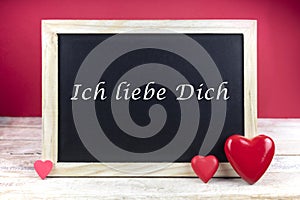Wooden blackboard with red hearts and written sentence in german Ich liebe dich, which means I love you, in red background