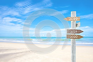 Wooden blank sign with text 2020 and 2021, Over Blurred blue sea and sand beach with cloudy blue sky