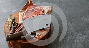 On a wooden block for meat a fresh raw tamahawk steak or a cowboy steak with a butcher`s chopping ax for meat, next to it is a