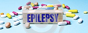 A wooden block with the word EPILEPSY stands on a blue background among multi-colored pills. Medical concept