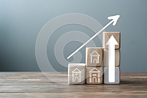 Wooden blocks with building illustrations ascend with white arrow on smooth surface against gray backdrop, symbolizing growth