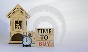 Wooden blocks form the words `time to buy` near miniature house. Black alarm clock. Beautiful white background. Business concept