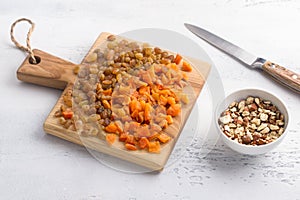 A wooden board with dried fruits - raisins and chopped dried apricots, a bowl with chopped hazelnuts and a knife on a light gray