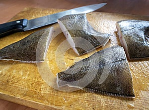 On a wooden board are large pieces of flounder and a knife. Raw fish is sliced and processed for eating. Cooking fish dishes