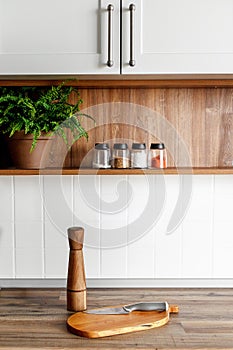 wooden board with pepper mill and knife on modern kitchen countertop and shelves with food ingredients and spices in glass jars.