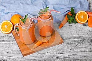 A wooden board with two mason jars with carrot smoothie, basil, oranges anf leaves of mint on a light background.
