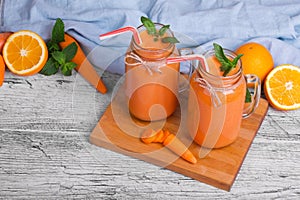 A wooden board with two mason jars with carrot smoothie, basil, oranges and leaves of mint on a light background.