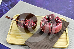 Wooden bowl containing wooden spoon and red pomegranate seeds. Pomegranate fruit open divided into five parts held together on a