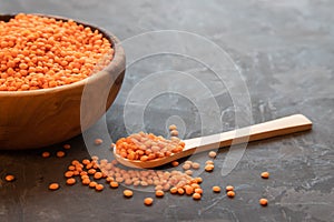 A wooden bowl full of raw red lentils and a wooden spoon filled with lentil grains next to a dark background. Place for text