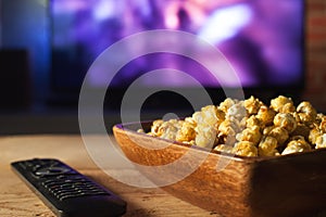 A wooden bowl of popcorn and remote control in the background the TV works. Evening cozy watching a movie or TV series at home