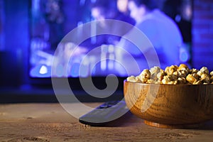 A wooden bowl of popcorn and remote control in the background the TV works. Evening cozy watching a movie or TV series at home