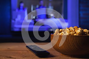 A wooden bowl of popcorn and remote control in the background the TV works. Evening cozy watching a movie or TV series at home