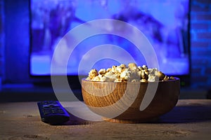 A wooden bowl of popcorn and remote control in the background the TV works. Evening cozy watching a movie or TV series at home