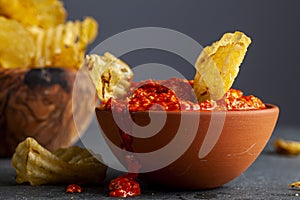 A wooden bowl of potato chips with a terra cotta bowl of salsa or hot pepper dipping sauce