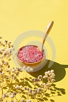 Wooden bowl with a spoon filled with pink bath sea salt. Beauty treatment for spa and wellness on yellow background.