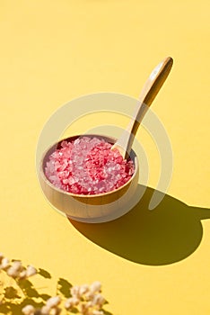Wooden bowl with a spoon filled with pink bath sea salt. Beauty treatment for spa and wellness on yellow background.