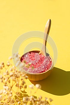 Wooden bowl with a spoon filled with pink bath sea salt. Beauty treatment for spa and wellness on yellow background.