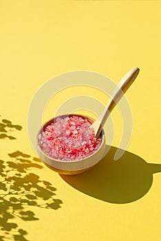 Wooden bowl with a spoon filled with pink bath sea salt. Beauty treatment for spa and wellness on yellow background.