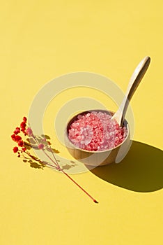 Wooden bowl with a spoon filled with pink bath sea salt. Beauty treatment for spa and wellness on yellow background.