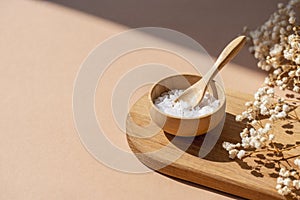 Wooden bowl with a spoon filled with white bath sea salt. Beauty treatment for spa and wellness on beige background.