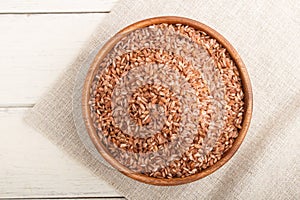 Wooden bowl with unpolished brown rice on a white wooden background. Top view, close up