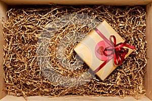 Wooden box with present wrapped in brown craft paper with red ribbon, filled with paper filler, top view