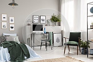 Wooden box under single metal bed with blue sheets and green blanket in trendy kid`s room with workspace