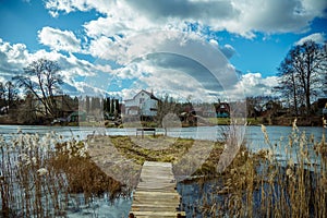 A wooden bridge leads from the shore to a small island on the lake. Fishing island on the background of clouds. A village and