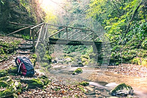 Wooden bridge over brook in autumn forest with backpack in foreground. Long exposure. Exploration concept, nature tracking, travel