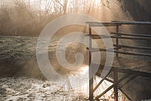 Wooden bridge over lake in early misty morning, fog over the water on sun rays