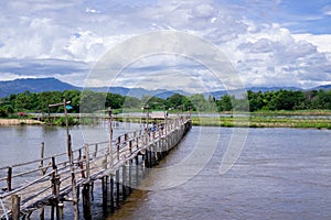 The wooden bridge over the river has a forested background. Mountains and blue skies